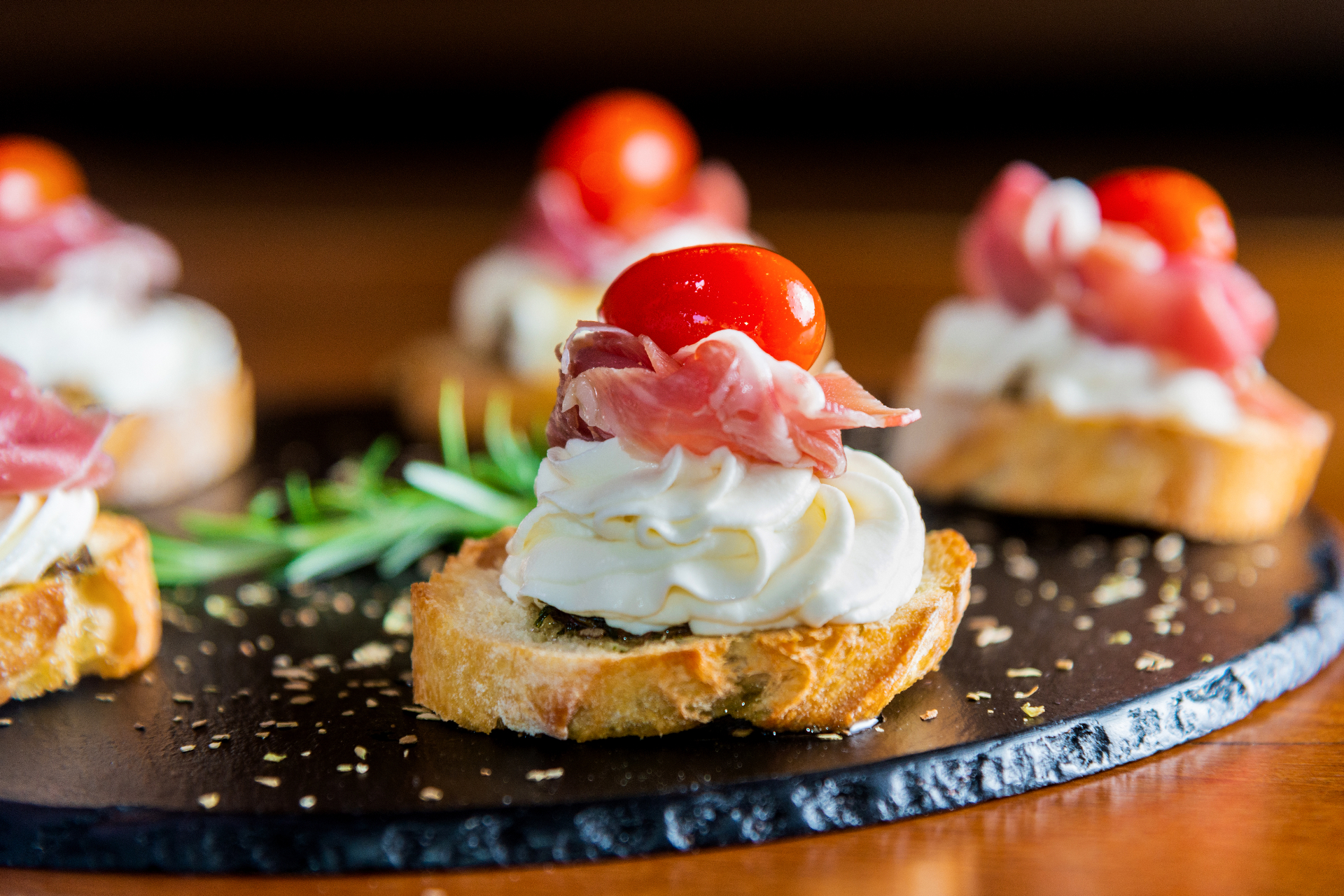 Bruschetta topped with creamy cheese, cured ham, and cherry tomato on a dark serving board