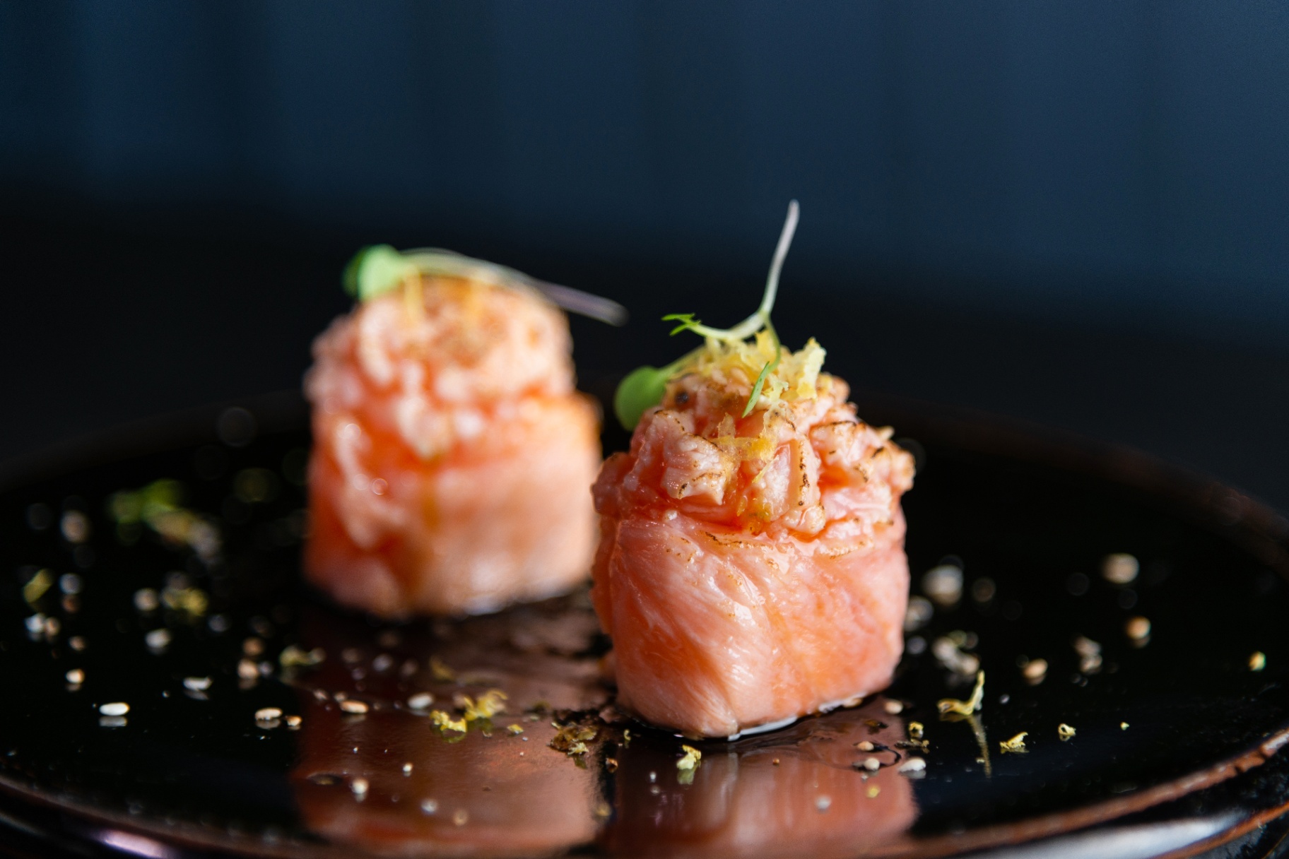 Two torched salmon bites topped with citrus zest on a dark ceramic plate