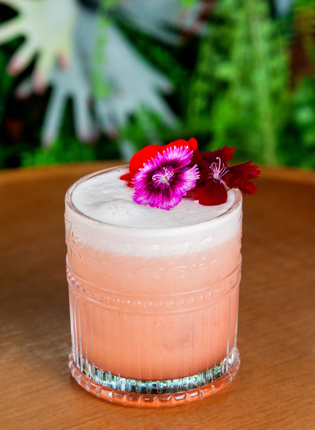 Pink foamy cocktail topped with edible flowers in a textured glass on wood